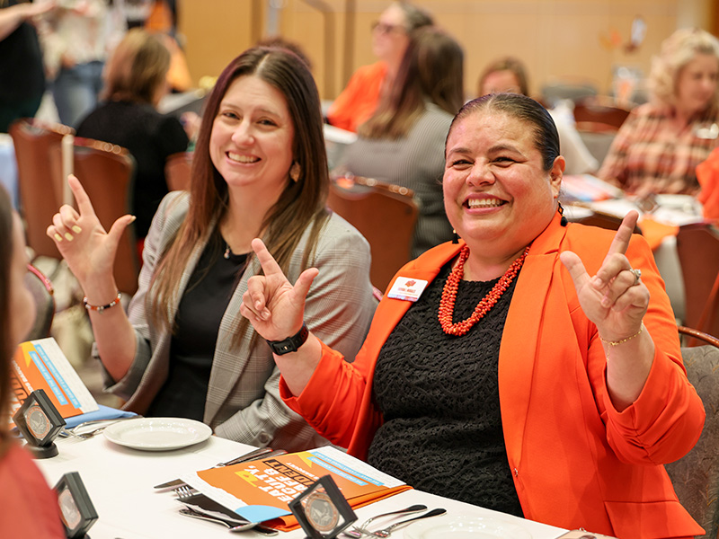 Two attendees sit at a table during a CEAT Faculty, Staff, and Student Awards Banquet, gesturing with their hands toward the camera, with event programs, place settings, and a medallion visible on the table.