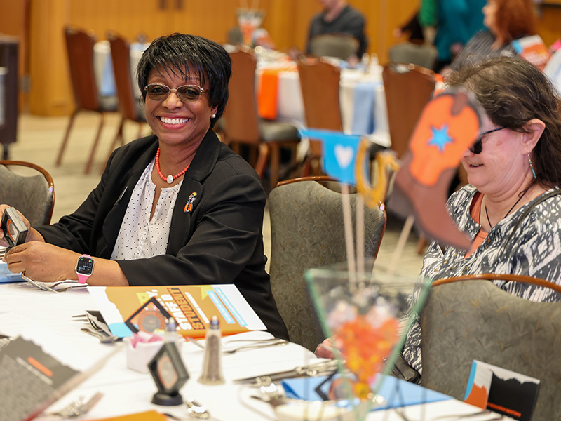 Two attendees are seated at a round table during a CEAT Faculty, Staff, and Student Awards Banquet, with printed programs, place settings, and a decorative centerpiece visible in a formal indoor event space.
