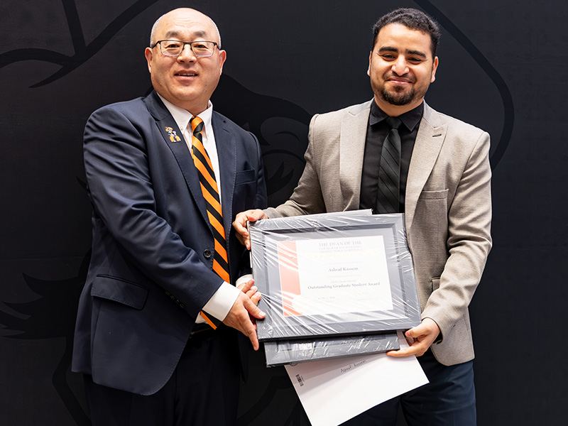 Two individuals stand in front of a dark backdrop holding a framed certificate together during a formal academic awards or recognition presentation.