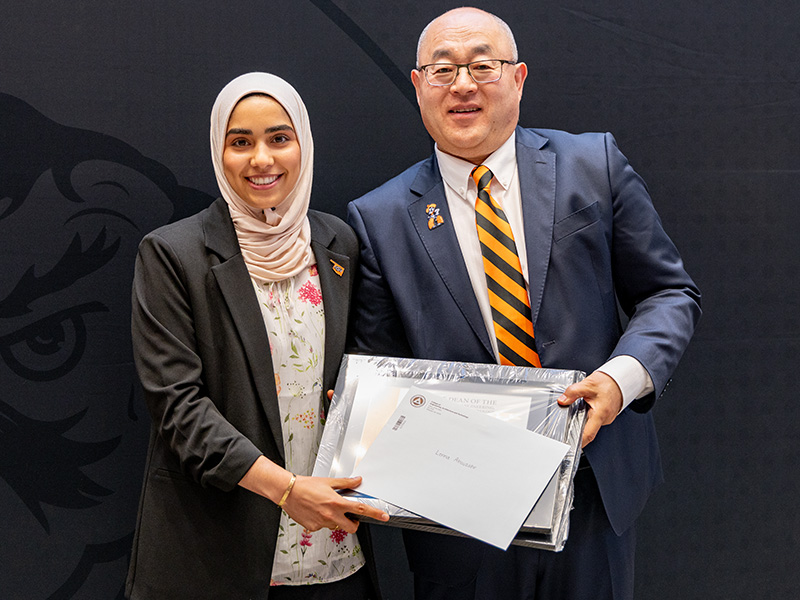 Two individuals stand in front of a dark backdrop holding a framed certificate together during a formal academic awards or recognition presentation.