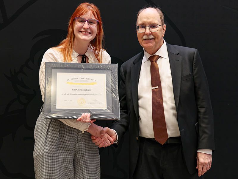 Two individuals stand in front of a dark backdrop shaking hands while one holds a framed certificate, documenting a formal academic or staff award recognition presentation.