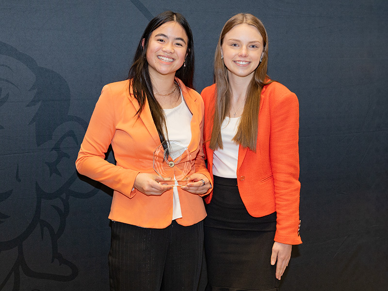 Two individuals stand side by side in front of a dark backdrop holding a clear glass award, documenting a formal student recognition or leadership award presentation.