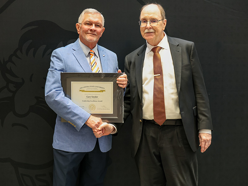 Two individuals stand in front of a dark backdrop shaking hands while one holds a framed certificate, documenting a formal academic or staff award recognition presentation.