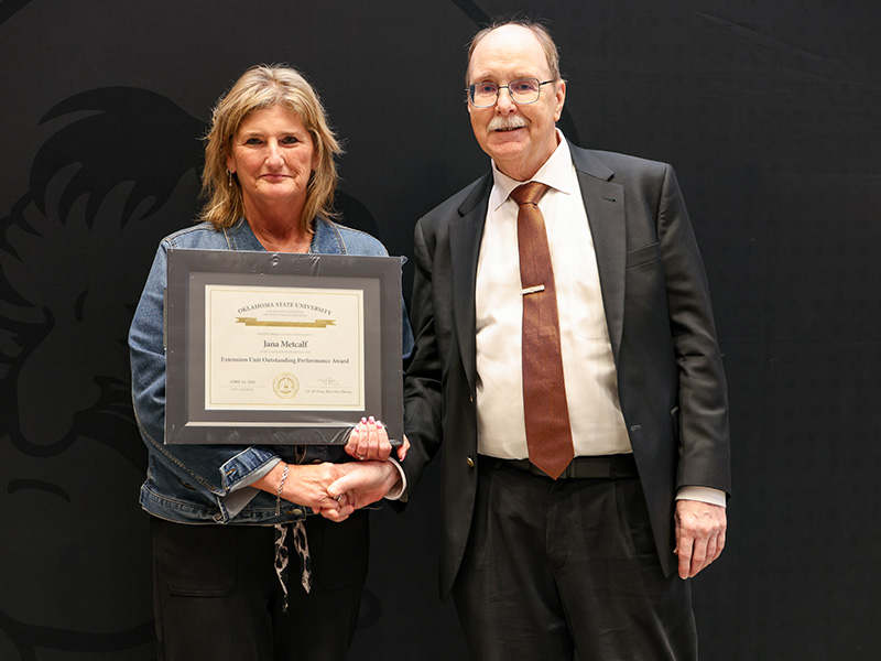 Two individuals stand in front of a dark backdrop during a formal award presentation, with one holding a framed certificate while they shake hands to mark an academic or staff recognition.