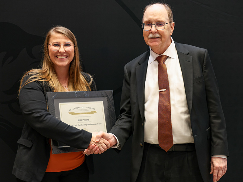 Two individuals stand in front of a dark backdrop shaking hands during a formal award presentation, with one holding a framed certificate to mark an academic or staff recognition.