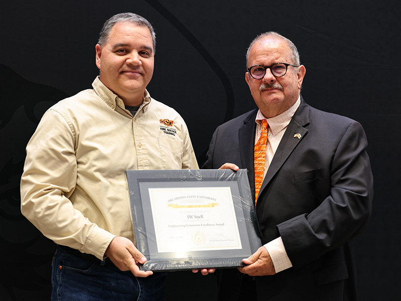 wo individuals stand in front of a dark backdrop holding a framed certificate together, documenting a formal academic or staff award recognition presentation.