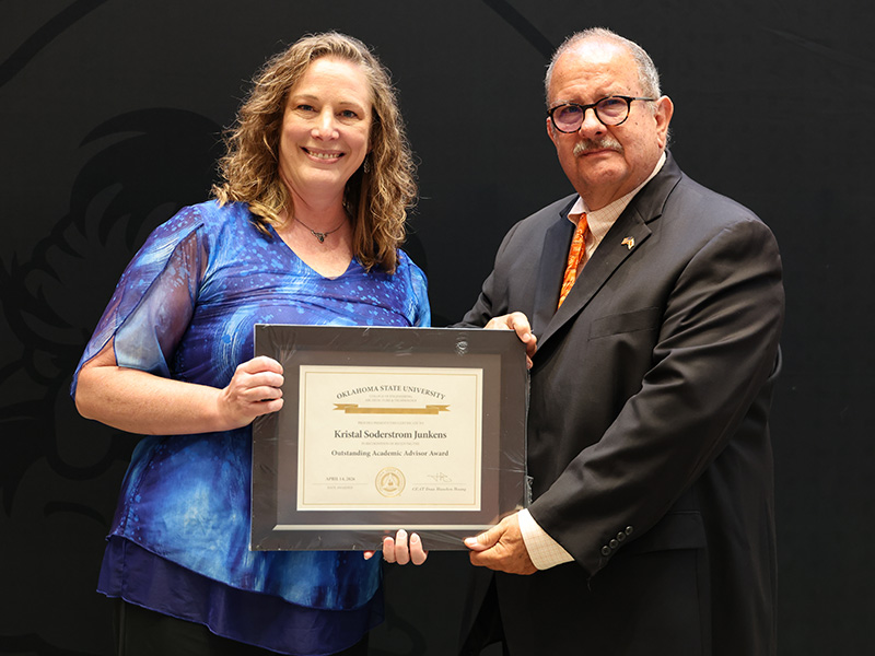 Two individuals stand in front of a dark backdrop holding a framed certificate together, documenting a formal academic or staff award recognition presentation.