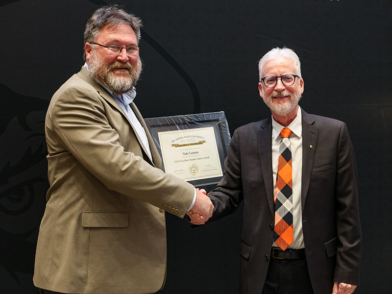 Two individuals stand in front of a dark backdrop shaking hands during a formal award presentation, with one holding a framed certificate to document an academic or staff recognition.