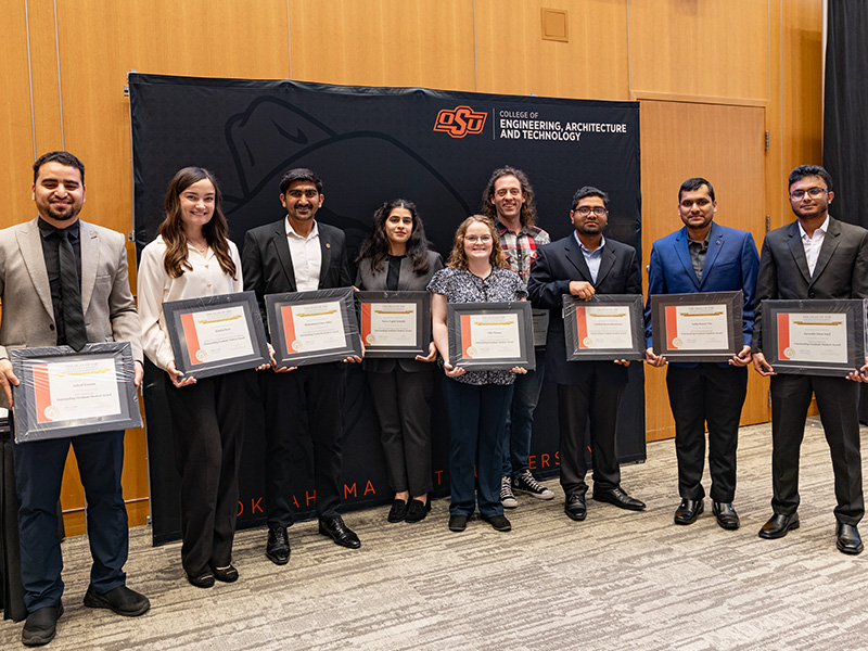 A group of individuals stand in front of an Oklahoma State University College of Engineering, Architecture and Technology backdrop, each holding a framed certificate to document recognition of outstanding graduate students during a formal awards presentation.