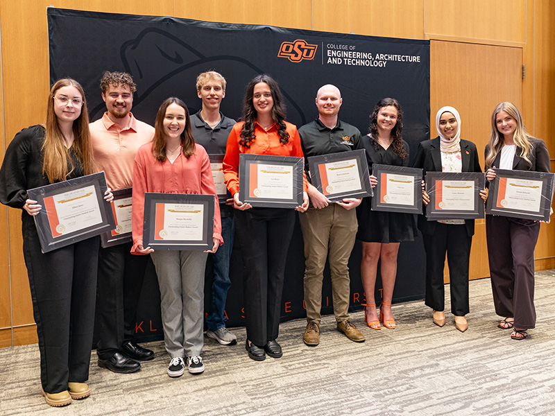 A group of individuals stand in front of an Oklahoma State University College of Engineering, Architecture and Technology backdrop holding framed certificates, documenting recognition of outstanding seniors during a formal awards presentation.
