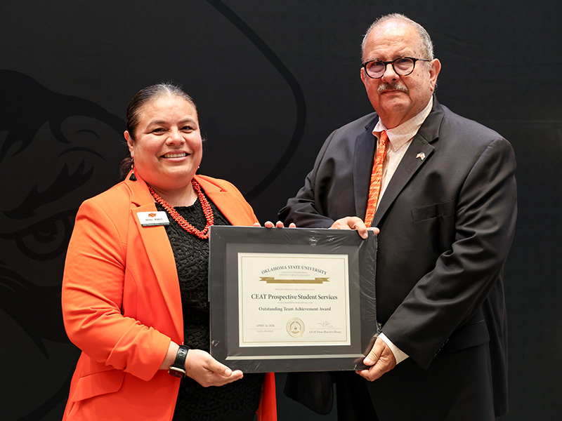 Two individuals stand in front of a dark backdrop holding a framed certificate together, documenting a formal team achievement award recognition for CEAT Prospective Student Services.