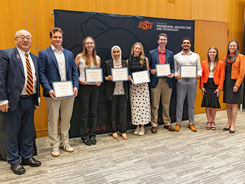 A group of individuals stand in front of an Oklahoma State University College of Engineering, Architecture and Technology backdrop holding framed certificates, documenting a formal student award or scholarship recognition presentation.