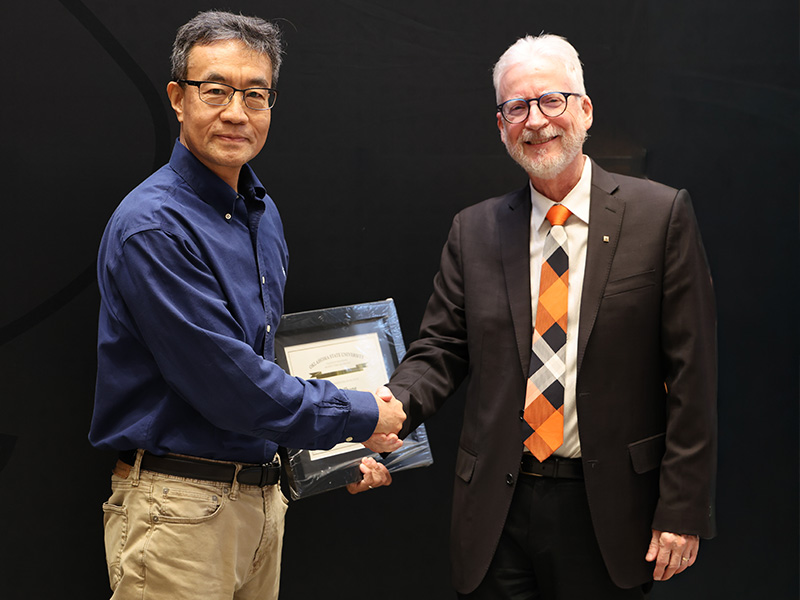 Two individuals stand in front of a dark backdrop shaking hands during a formal award presentation, with one holding a framed certificate to document an academic or staff recognition.