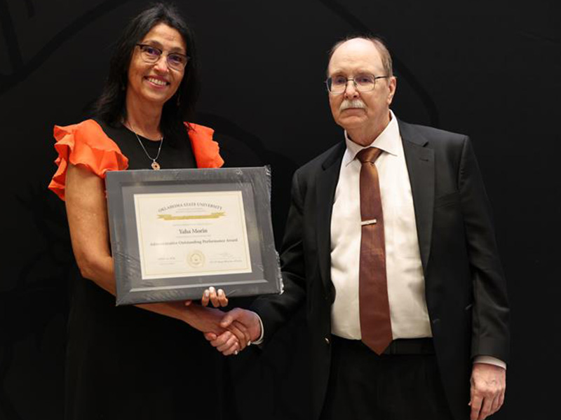 Two individuals stand in front of a dark backdrop during a formal award presentation, with one holding a framed certificate while they clasp hands to document an academic or staff recognition.