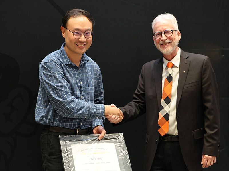 Two individuals stand in front of a dark backdrop shaking hands during a formal award presentation, with one holding a framed certificate to document an academic or staff recognition.