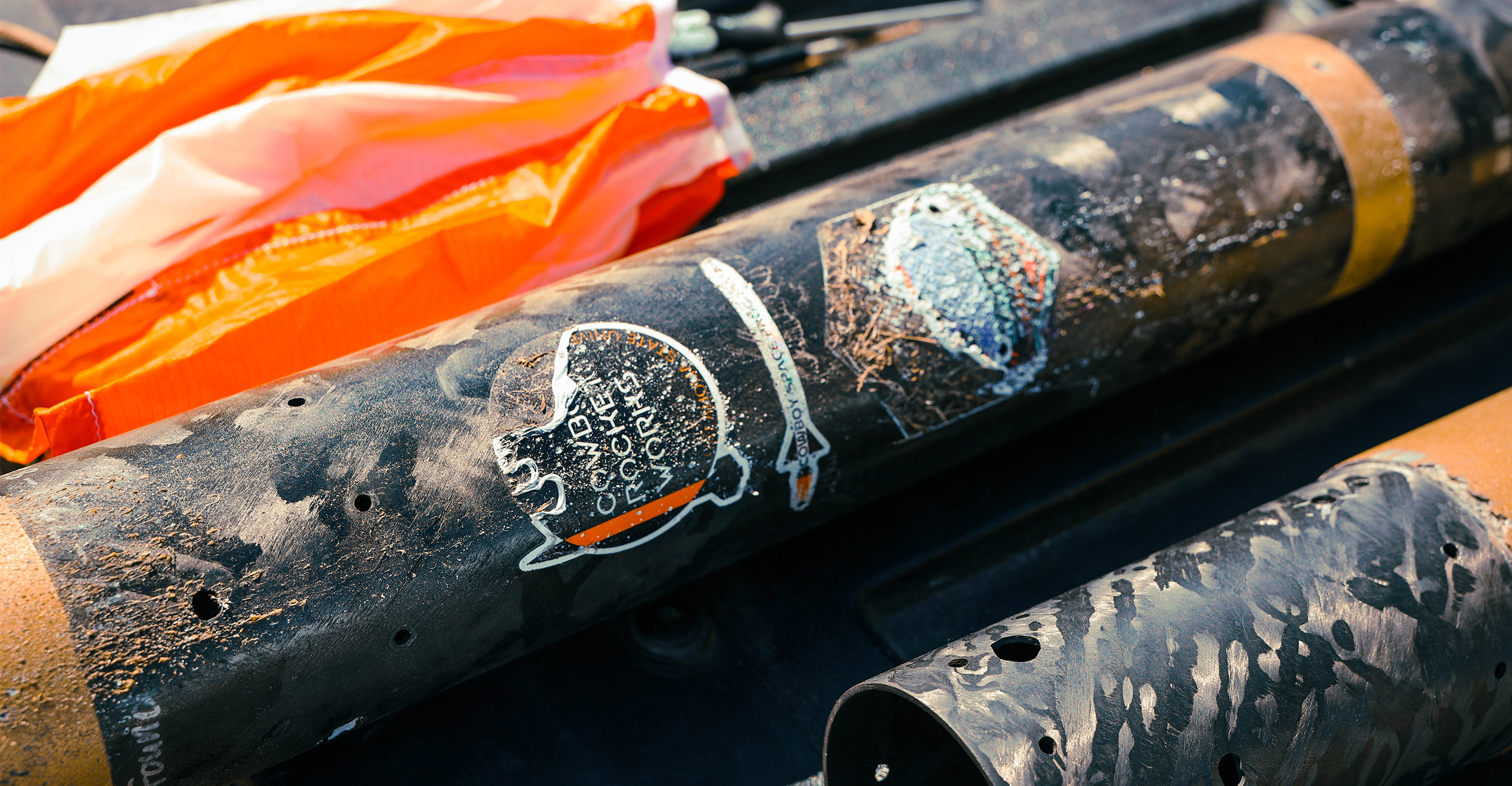 Close-up of worn cylindrical metal equipment with peeling labels and drilled holes, resting alongside an orange safety fabric, highlighting used industrial or drilling components.
