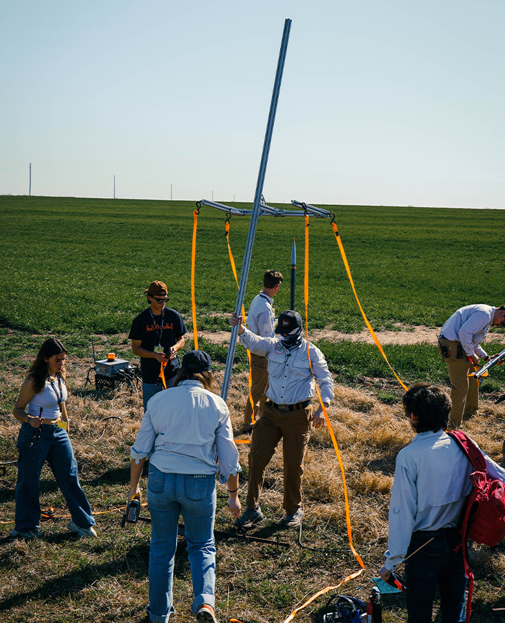 A group of people in an open field assemble and stabilize a vertical metal launch rail with straps while preparing model rocketry equipment for an outdoor launch.