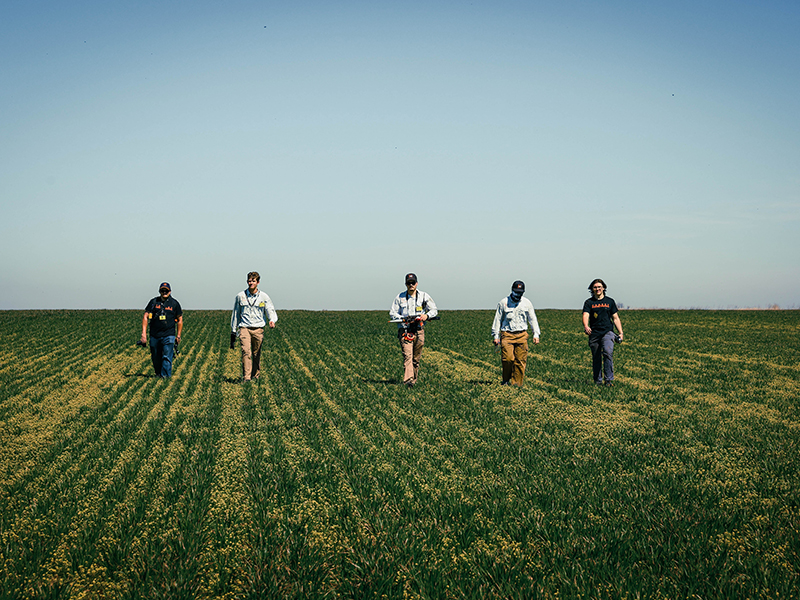 Several people walk across a wide, open grassy field in a horizontal line, carrying small equipment and wearing casual outdoor clothing under a clear sky.
