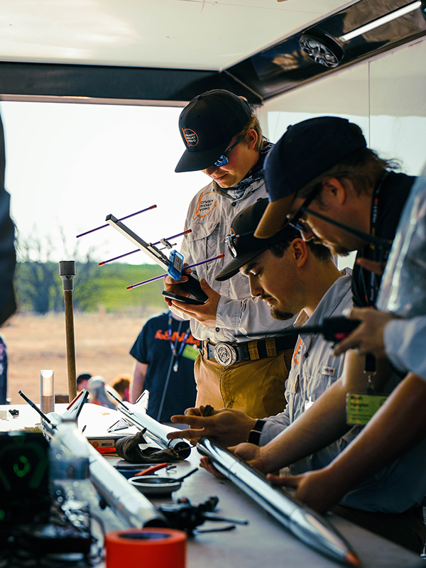 Several people work together at a table inside a mobile workspace, closely inspecting and assembling long, cylindrical rocket components using tools and electronic equipment during a rocketry preparation activity.