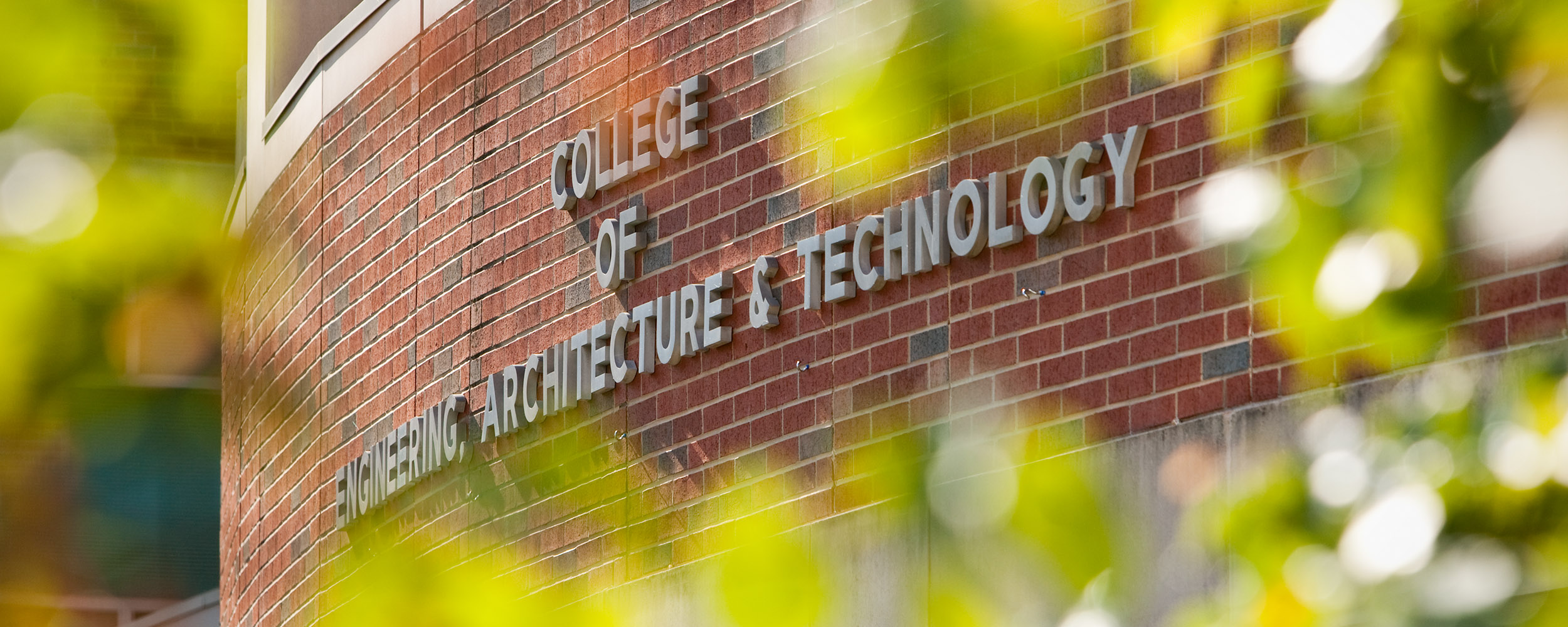 A curved brick building exterior displays raised lettering reading “College of Engineering, Architecture & Technology,” partially framed by out-of-focus green leaves in the foreground, identifying the academic facility on campus.