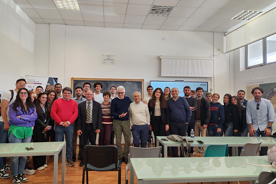A large group of people pose together inside a classroom, standing in front of desks and a chalkboard with presentation screens, indicating a group photo taken after a lecture, workshop, or academic event.