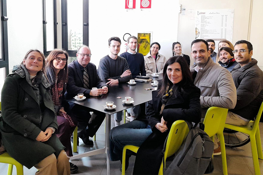 A group of people sit closely around a table inside a café or break room, with small cups of coffee on the table, posing together during an informal social or professional gathering.