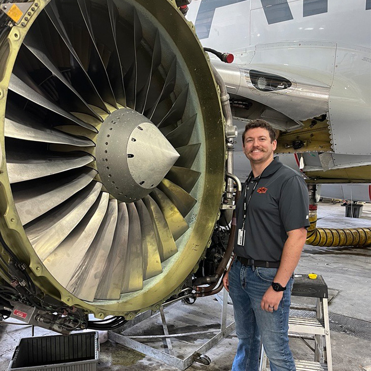 A person wearing an OSU-branded polo stands beside a large exposed aircraft jet engine inside a maintenance hangar, highlighting aviation training or engineering work.