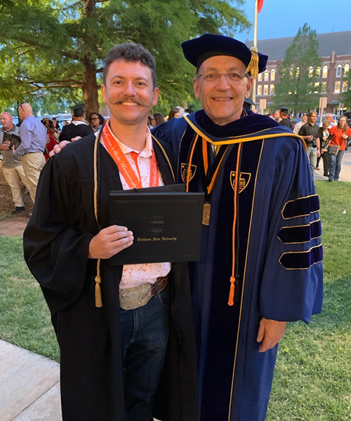 Two people stand outdoors at a graduation event, one wearing a cap and gown and holding an Oklahoma State University diploma cover while posing beside another person in academic regalia, with a crowd and campus buildings in the background.