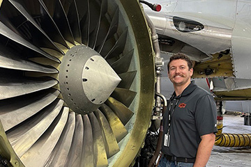 A person wearing an OSU-branded polo stands beside a large exposed aircraft jet engine inside a maintenance hangar, highlighting aviation training or engineering work.