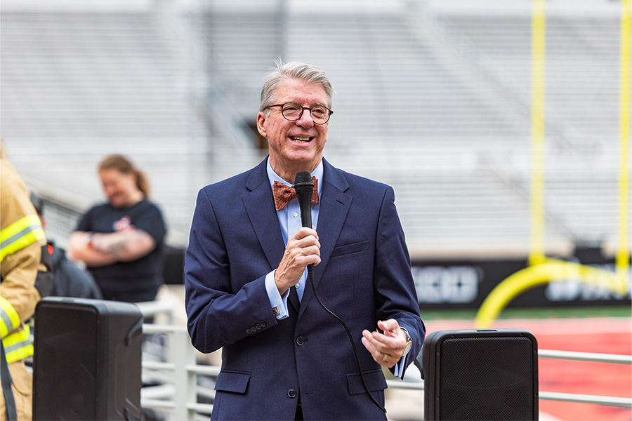 A gray-haired man wearing glasses, a navy suit, light blue shirt, and orange bow tie stands in the foreground, speaking into a handheld microphone