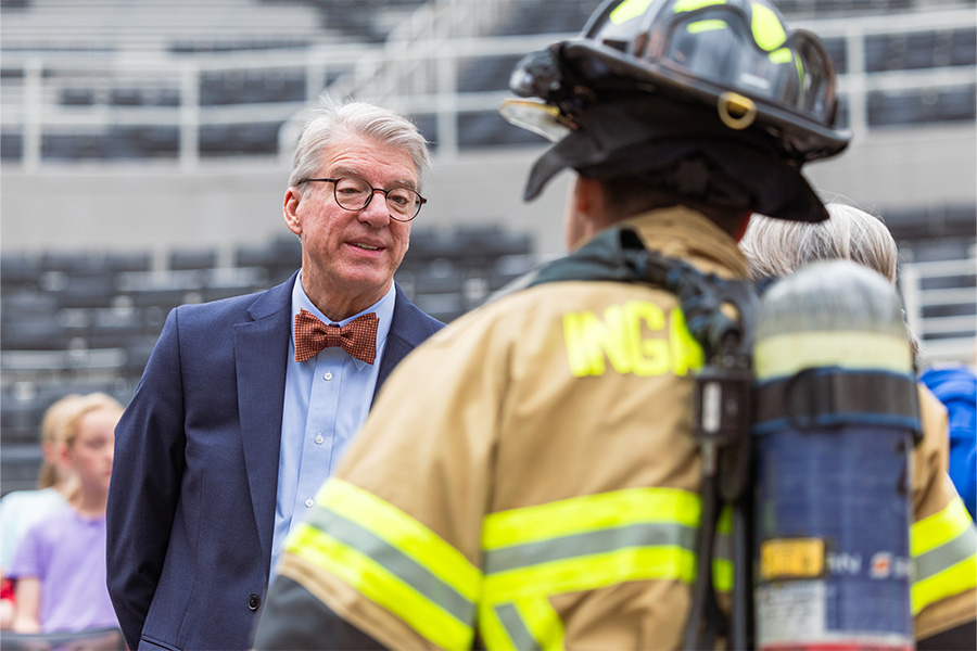 A close-up outdoor scene shows an older man in a navy suit, light blue shirt, and orange bow tie speaking with a firefighter.