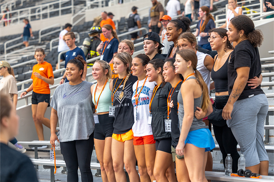 A group of young women and girls pose together on indoor bleachers at a sporting event.