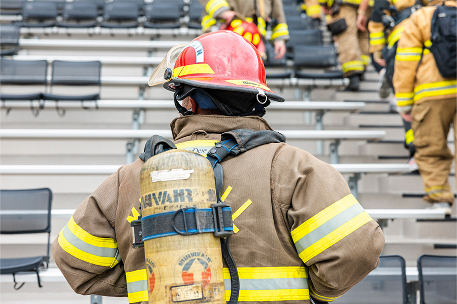 A firefighter is shown from behind in the foreground, wearing a brown protective coat with bright yellow reflective stripes, a red helmet, and a breathing apparatus secured to their back