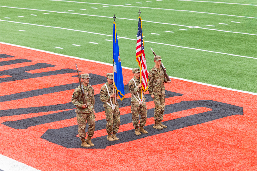 Four uniformed service members stand in formation on an orange football end zone.