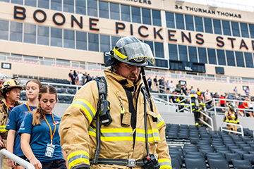 A firefighter in tan protective gear and a helmet walks through the foreground of a football stadium.