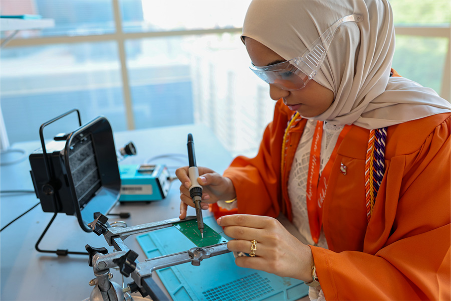 A person wearing safety goggles works at a lab workstation, using a soldering iron to assemble or repair an electronic circuit board secured in a holder.