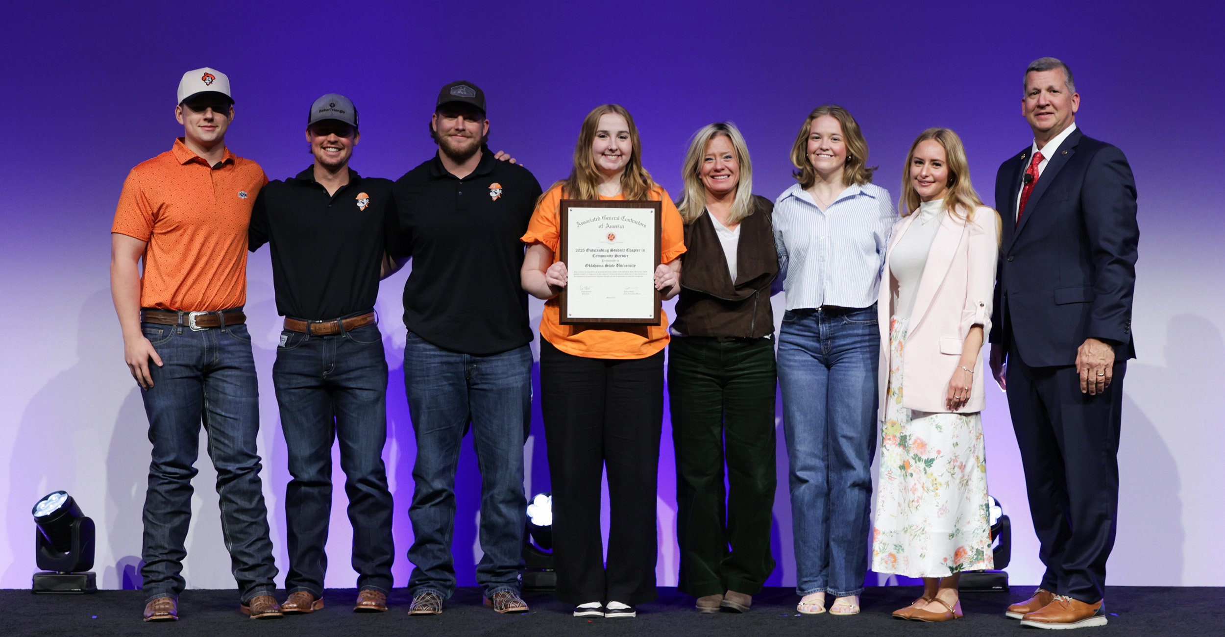 Group of eight people standing on a stage with purple lighting, centered around one person holding a certificate, posed for a formal award or recognition photo.