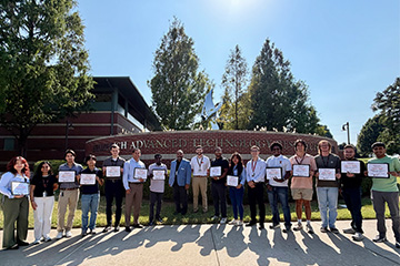 Group of students and faculty members standing outdoors in front of an engineering building, holding certificates and posing for a recognition photo.
