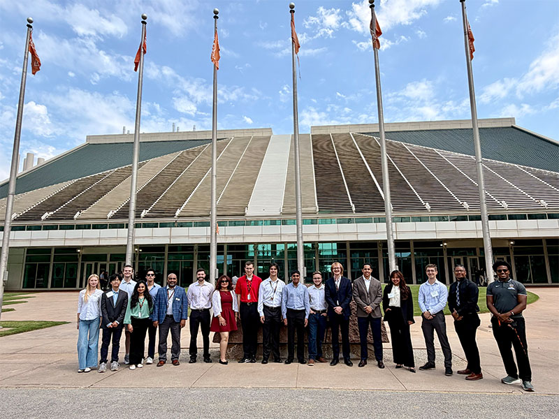 A group of students, faculty, and researchers pose for a photo in front of a large Oklahoma State University building. Several OSU flags are displayed on tall flagpoles above the group against a blue sky with scattered clouds.