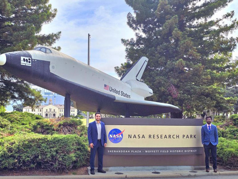 Two professionally dressed individuals stand beside a sign for NASA Research Park beneath a space shuttle display at Moffett Field in California. Trees and landscaped grounds surround the outdoor exhibit.
