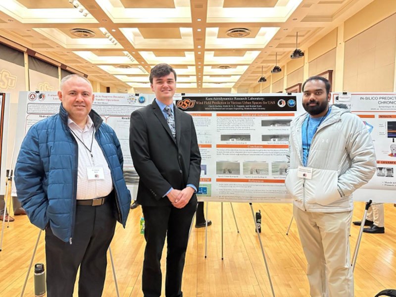 Three attendees stand in front of a research poster during an academic poster session at Oklahoma State University. The poster is displayed on an easel in a large conference hall with additional research displays visible in the background.