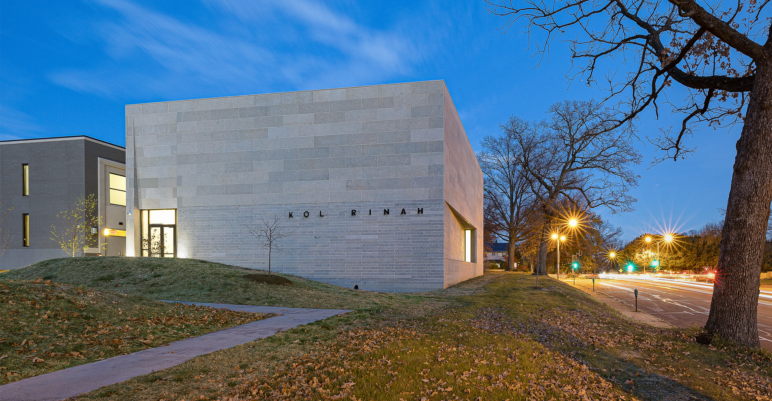 A modern light-colored building labeled “KOLRINAH” sits along a tree-lined street at dusk, with illuminated windows and passing car lights under a blue evening sky.
