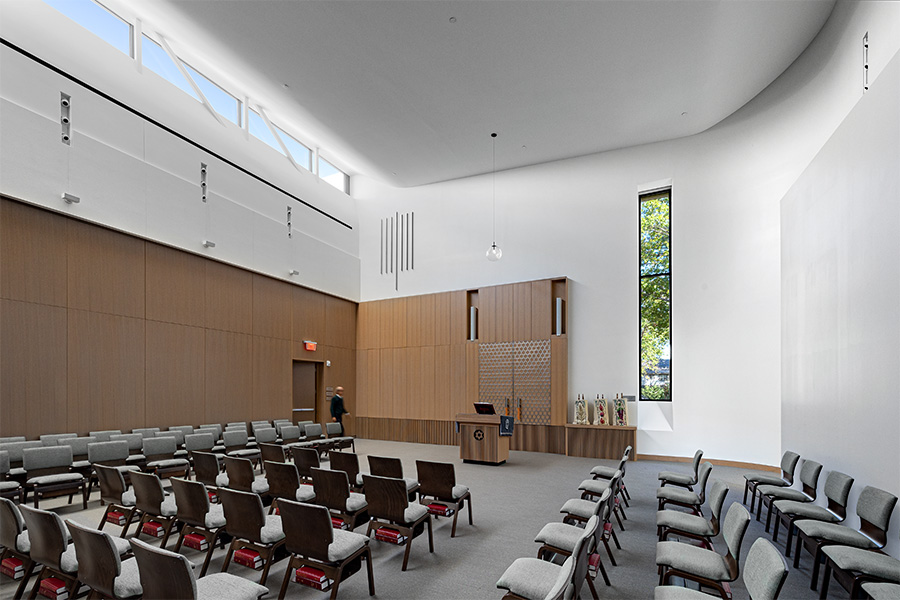 A bright, modern interior of a chapel-like space features rows of chairs facing a small podium and decorative wall, with tall windows and high ceilings allowing natural light to fill the room.