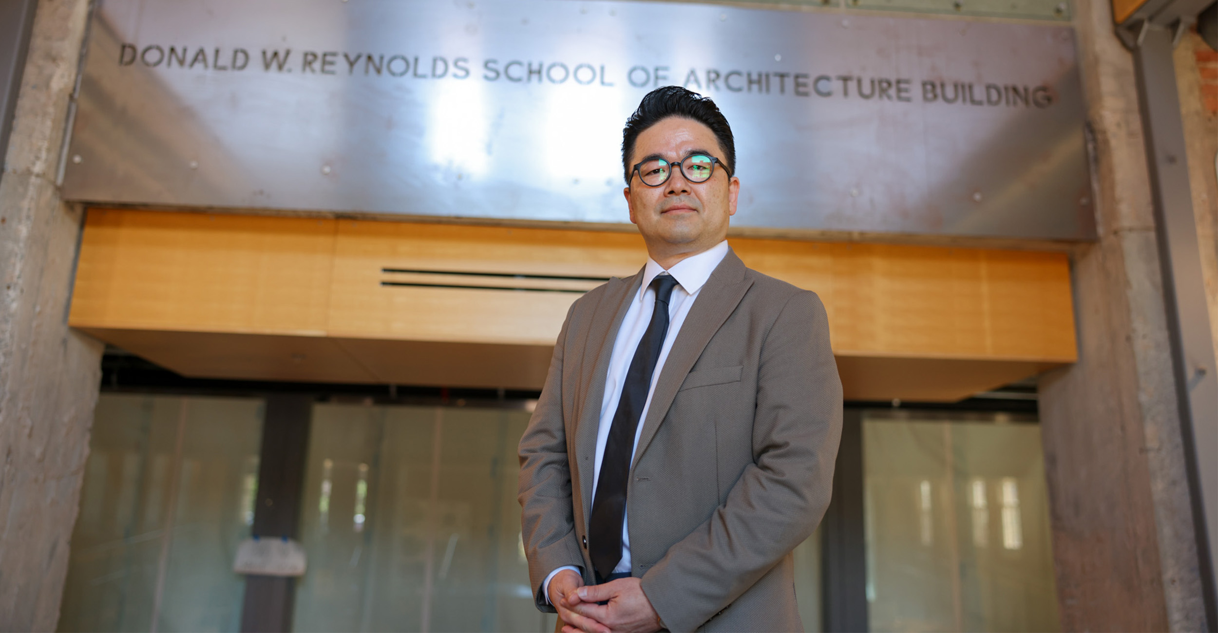 A person in a suit and glasses stands indoors beneath a sign reading “Donald W. Reynolds School of Architecture Building,” posed for a professional portrait in front of a modern interior entryway.