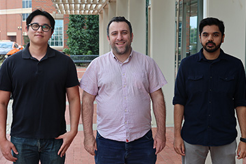 Three men standing side by side outdoors on a brick walkway, near a building with large windows and a covered walkway.