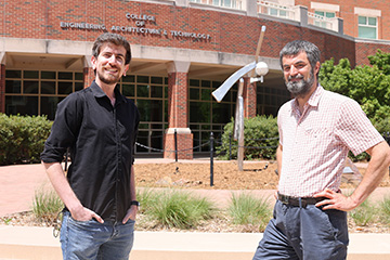 Two men smiling outdoors in front of the College of Engineering, Architecture, and Technology building, surrounded by greenery and sculptures.