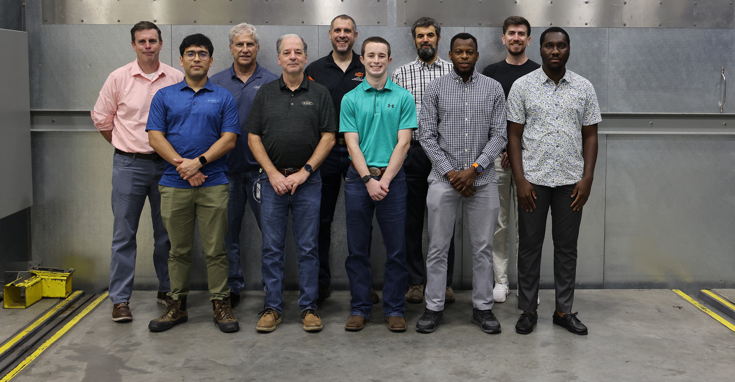 A group of ten adults stands in two rows for a posed photograph in an industrial-looking room. The group is arranged against a gray metal wall with riveted panels, and the concrete floor has yellow-and-black painted markings and equipment tracks.