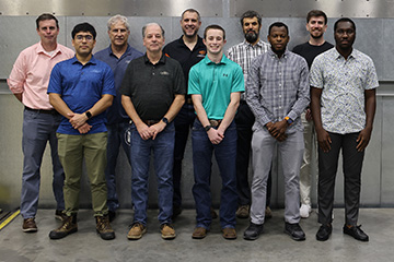A group of ten adults stands in two rows for a posed photograph in an industrial-looking room. The group is arranged against a gray metal wall with riveted panels, and the concrete floor has yellow-and-black painted markings and equipment tracks.