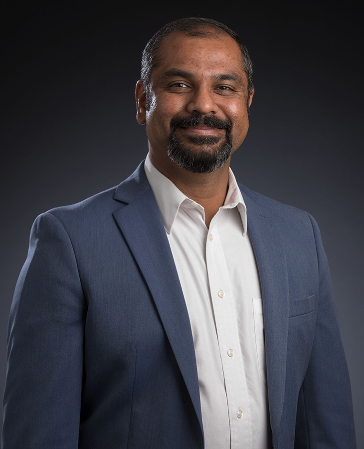 Studio portrait of a person wearing a blue blazer and white shirt, standing against a dark gradient background.
