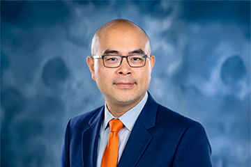 A man in a navy blue suit and orange tie poses for a professional headshot against a blue and white swirled backdrop.
